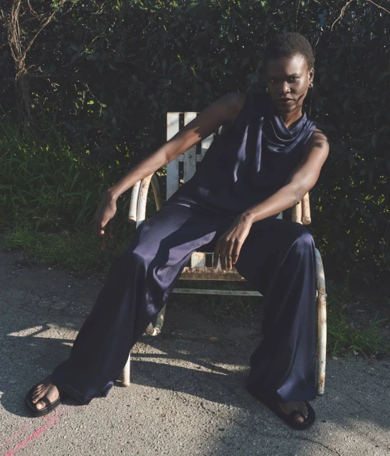 A fashionable Black woman in a navy jumpsuit sits on a white chair outdoors, posing in natural light with a serious facial expression.