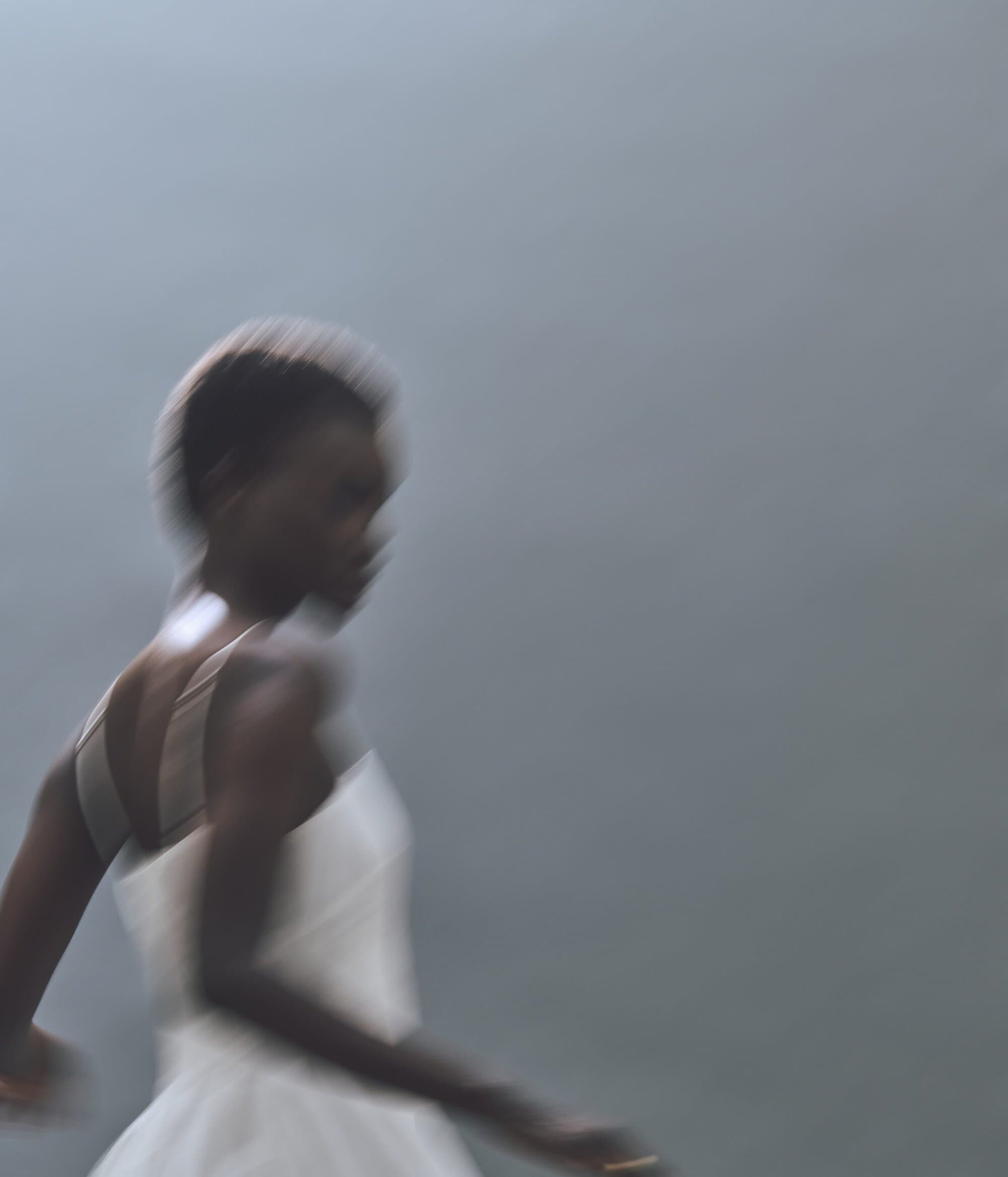 Motion blur of a Black woman in a light dress against a grey background, creating an abstract and dynamic fashion image.