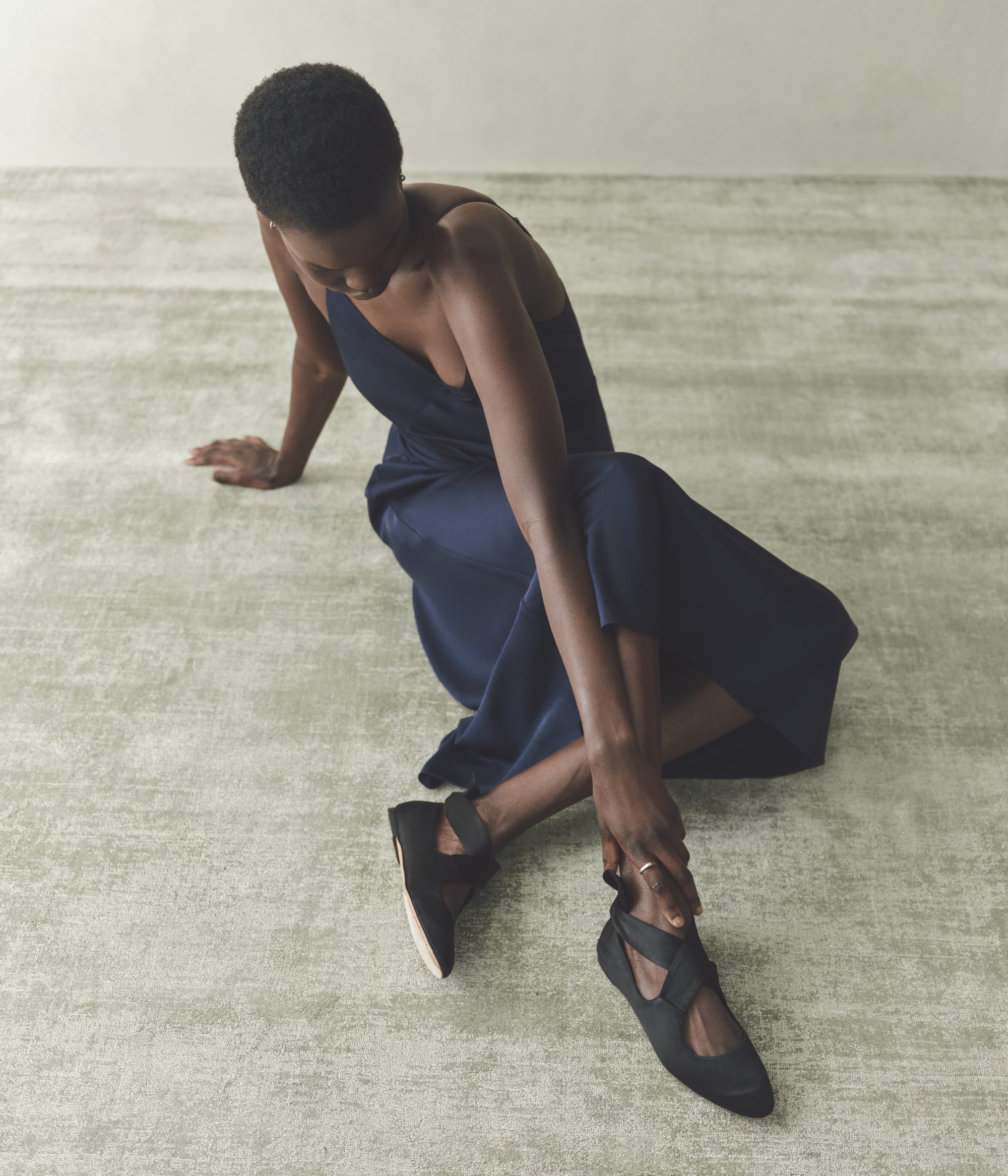 Woman in a navy slip dress and black ballet flats sitting on a gray textured floor in a brightly lit neutral setting.