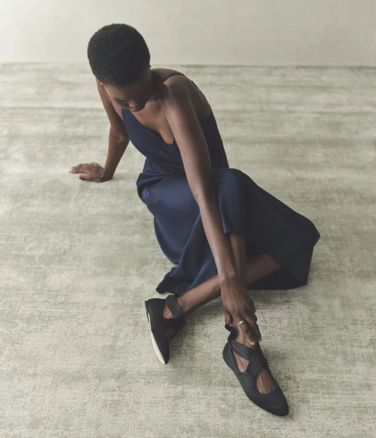 Woman in a navy slip dress and black ballet flats sitting on a gray textured floor in a brightly lit neutral setting.
