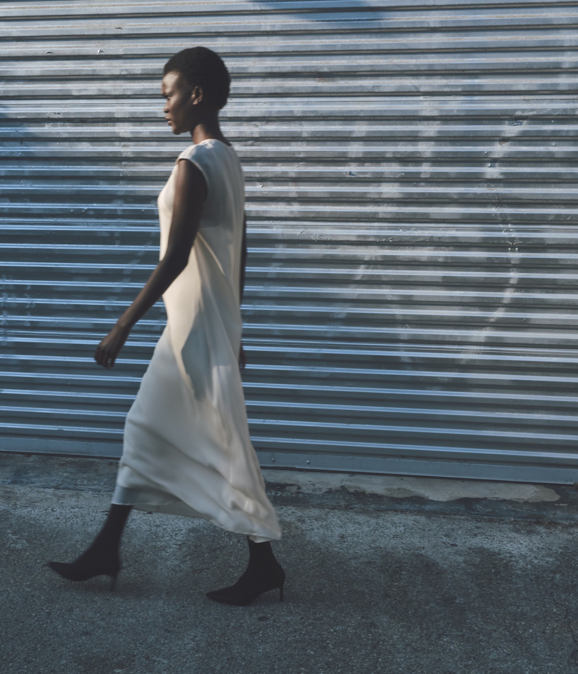Elegant woman in a flowing dress walks against a textured metal wall. Soft natural lighting casts shadows. Low angle, full shot.