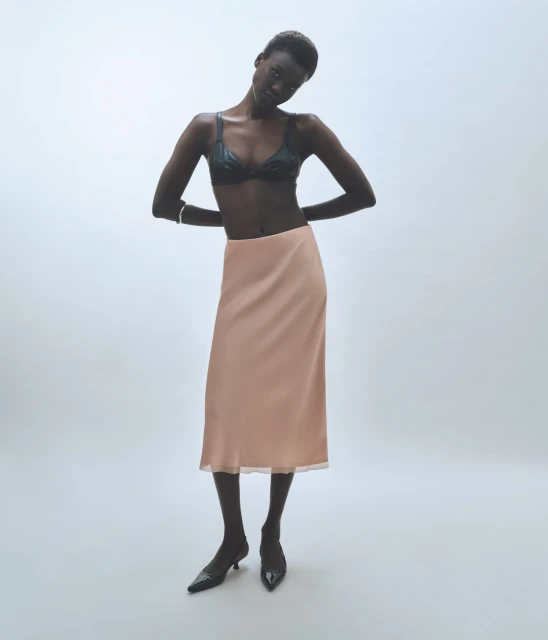 An African-American woman poses in a bra and skirt against a white background, showcasing a fashion-forward, minimalist look.
