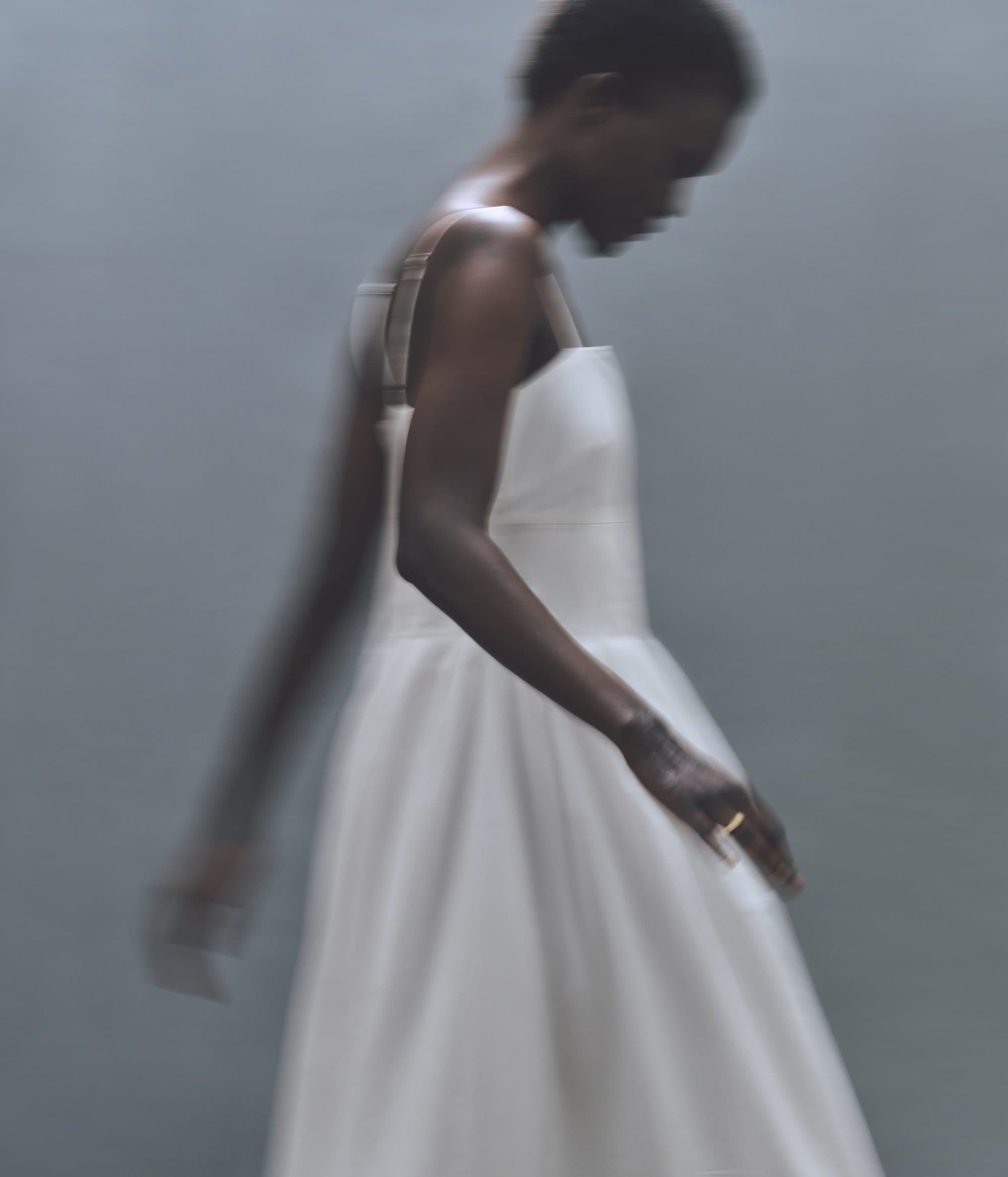 Woman in a white dress with a square neckline stands in a blurred view of a photo studio with a soft light and a simple backdrop.