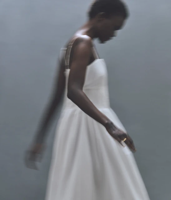 Woman in a white dress with a square neckline stands in a blurred view of a photo studio with a soft light and a simple backdrop.