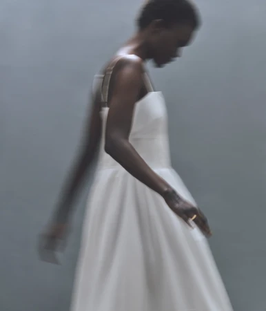 Woman in a white dress with a square neckline stands in a blurred view of a photo studio with a soft light and a simple backdrop.