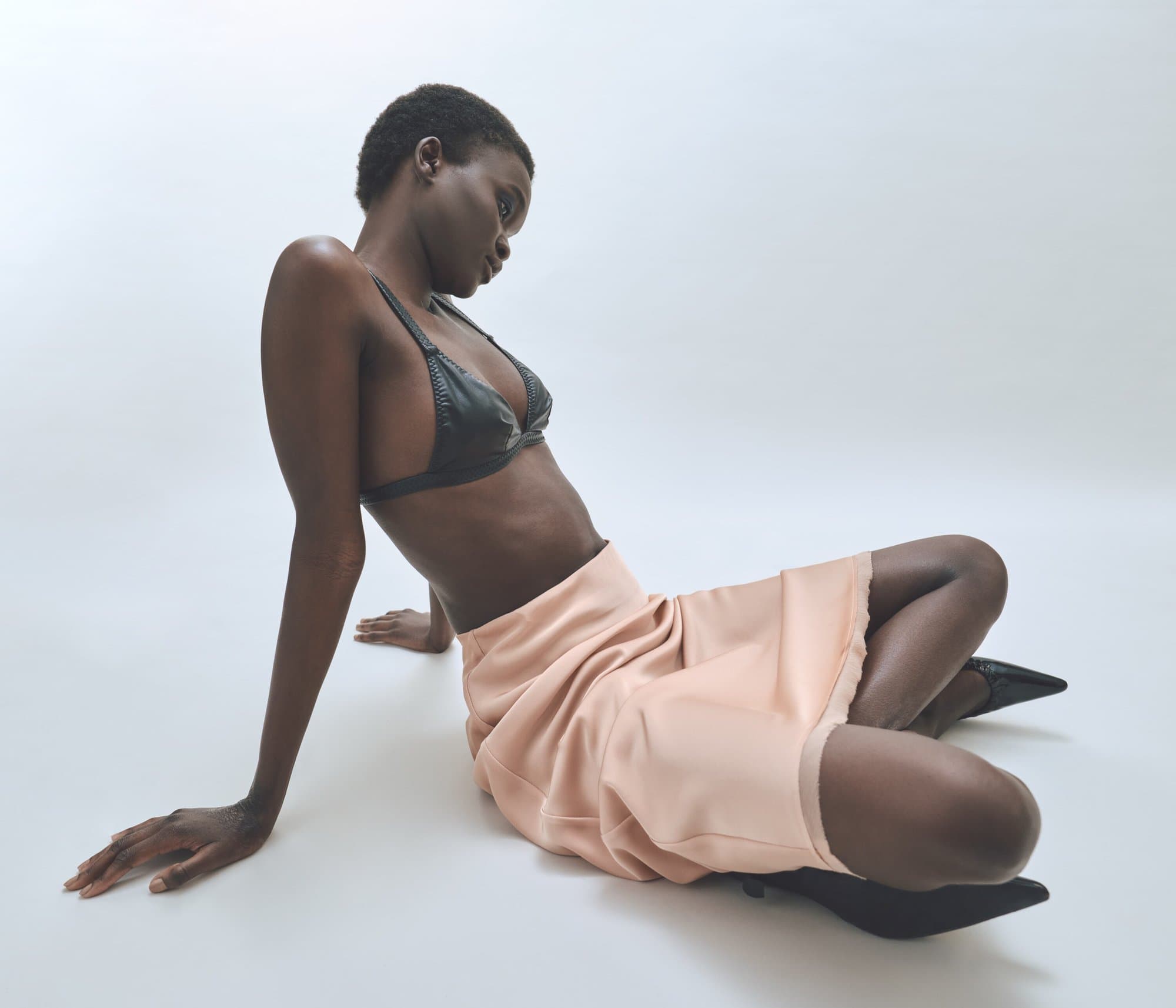 Dark-skinned woman in a black bra and peach-colored skirt poses on a white background in a fashion-style full shot with studio lighting.