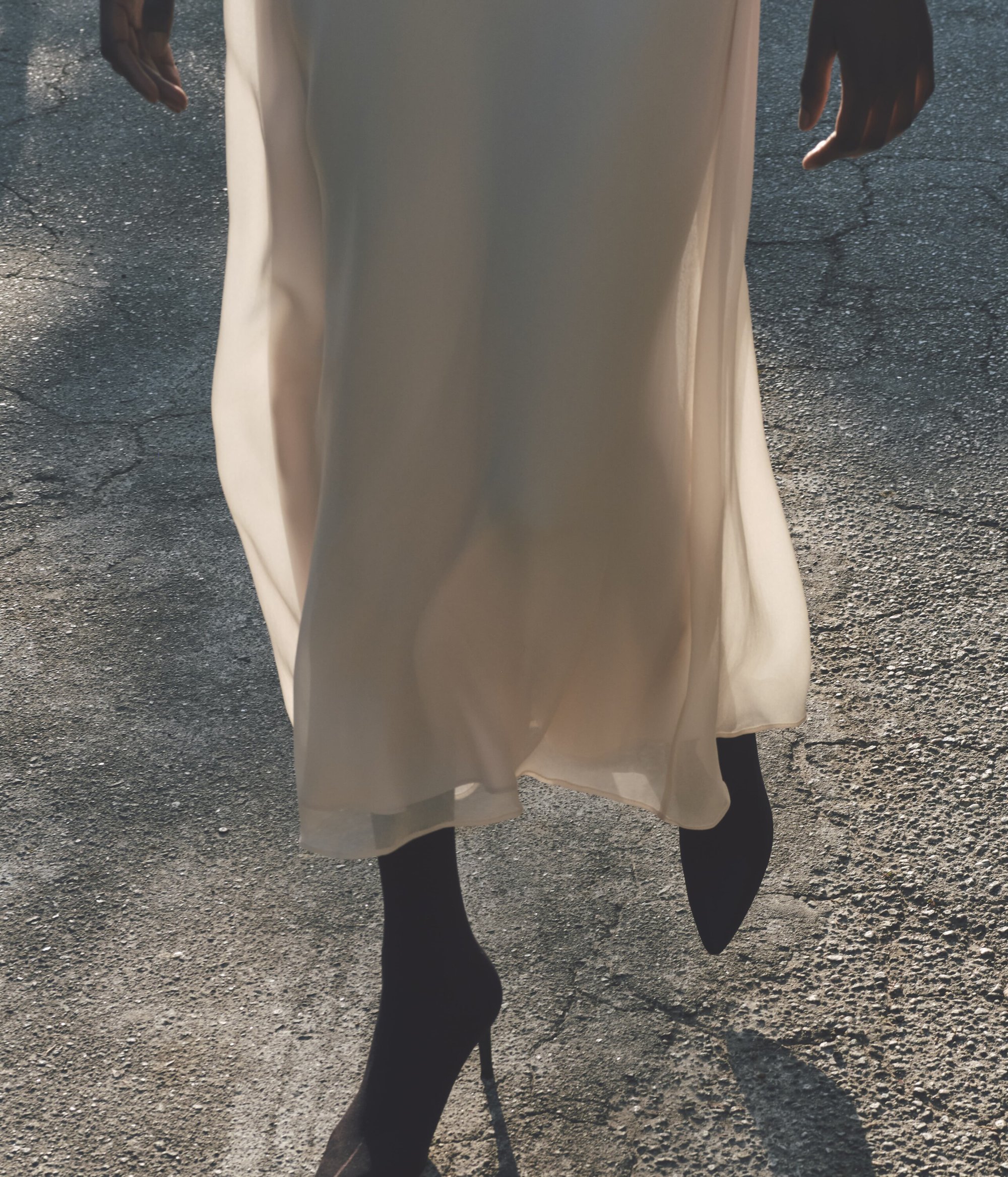Fashion shot of a dark-skinned woman walking in a cream-colored dress and black high heels on an asphalt road, capturing a blend of shadow and light.
