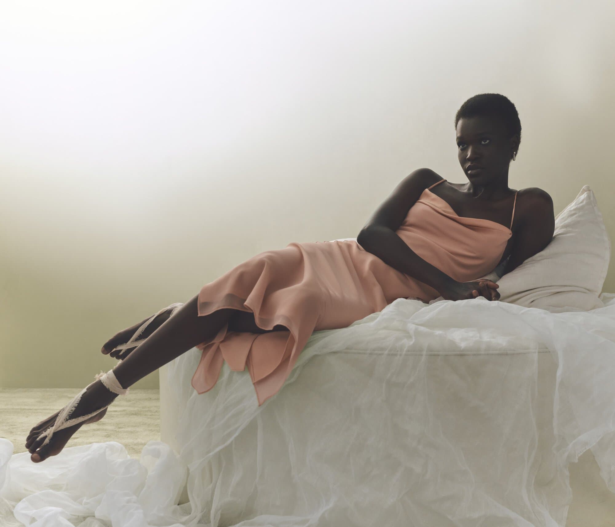 Elegant Black woman in a peach gown lies on a white bed. Soft light and fabrics create a serene fashion editorial look indoors.