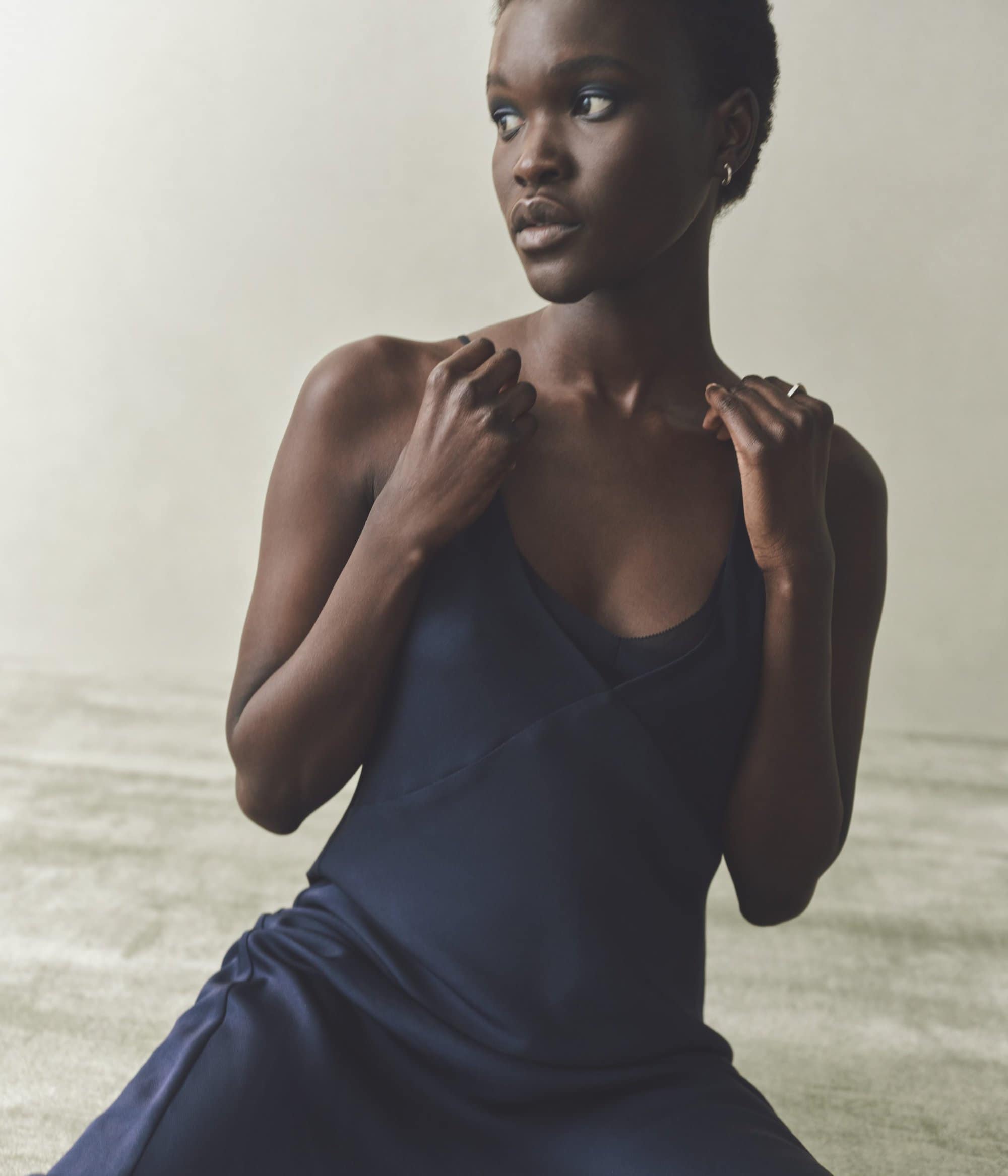 Portrait of a dark-skinned woman in a navy dress. She is indoors on a neutral background, looking off-camera with a subtle expression.