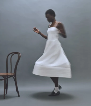 Black woman in a white dress twirling next to a wooden chair against a grey seamless background
