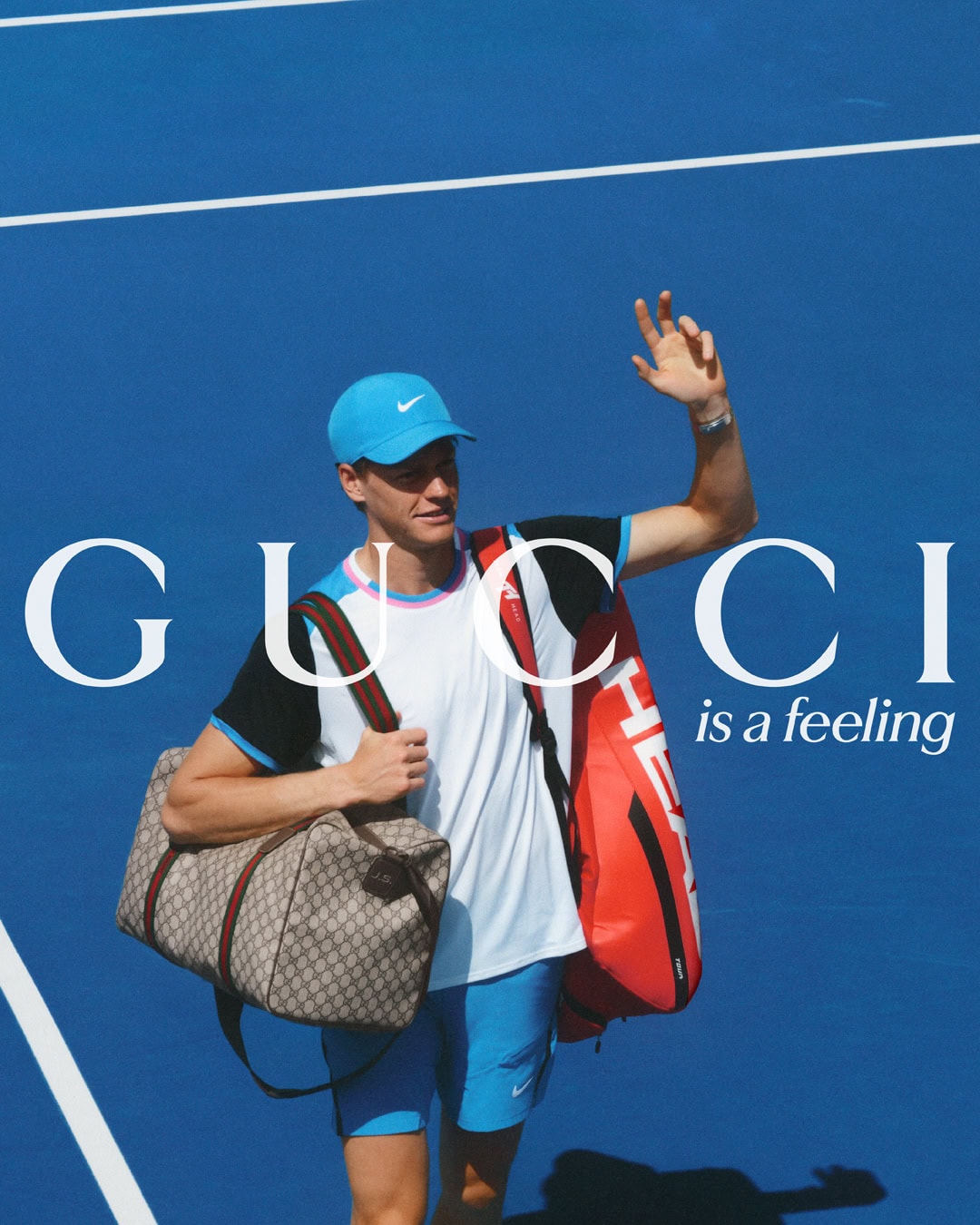 A male tennis player in athletic wear, wearing a blue cap, carries a tennis bag and Gucci duffel on a blue tennis court surface.