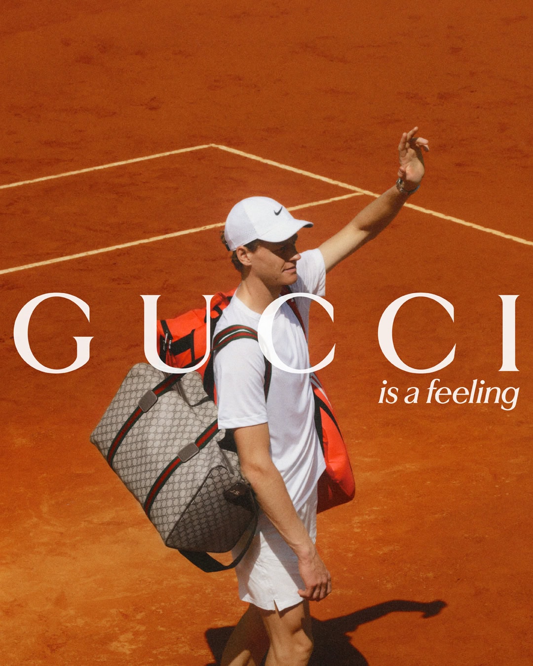 A male tennis player stands on a clay court with a duffel bag and gear, advertising Gucci, with text that reads 'Gucci is a feeling'.