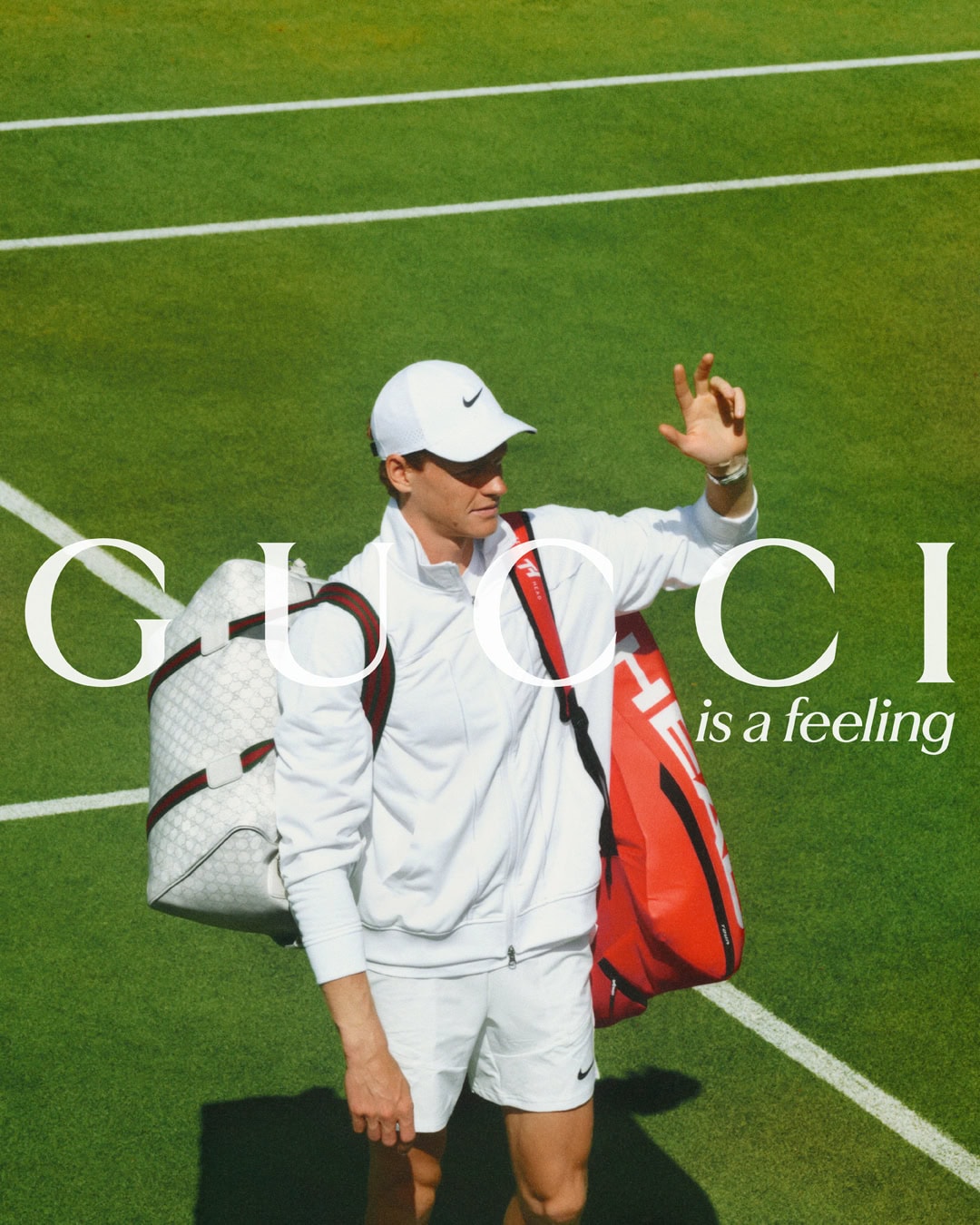 Male tennis player on court with Gucci bag and red HEAD tennis bag, wearing white Nike hat and outfit, with Gucci logo.