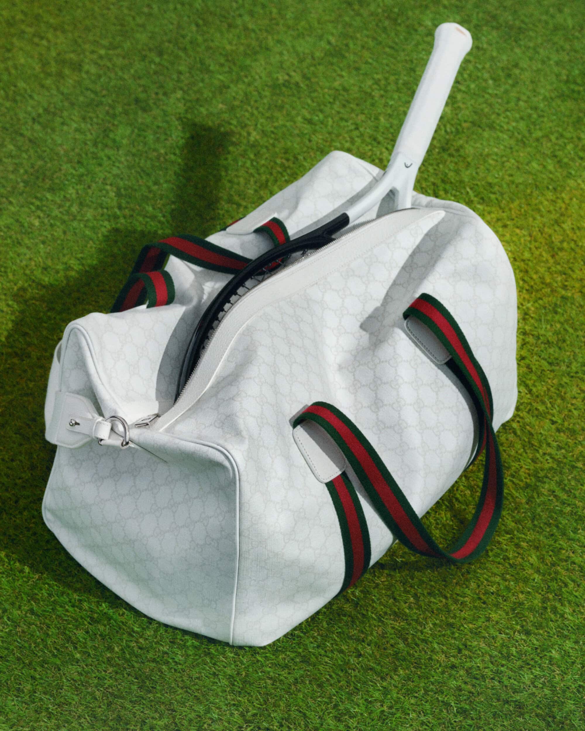 White Gucci duffel bag with green and red stripes and a tennis racket inside, resting on artificial grass in a studio setting.