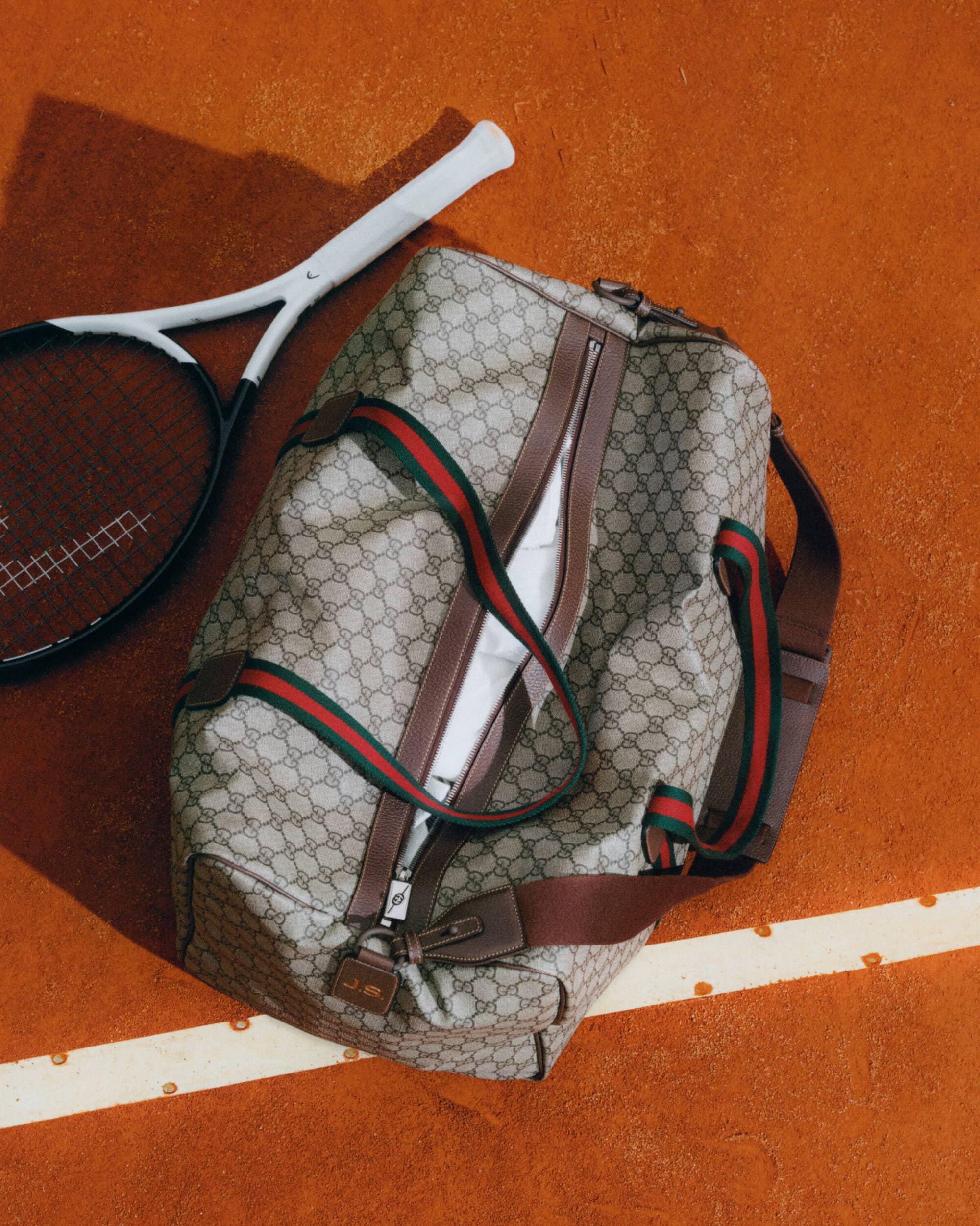 Tan Gucci duffel bag and white tennis racket on a red tennis court. A sporty image with luxury fashion and an athletic lifestyle.