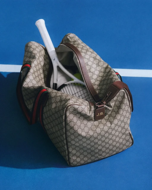 Tan Gucci duffle bag with tennis racquet inside on a blue tennis court. The bag has brown leather trim and red and green stripes.