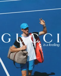 Jannik Sinner waving on a blue tennis court carrying a monogram duffle bag for a Gucci campaign