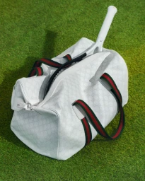 White leather duffle bag with green and red striped straps resting on green turf grass with a tennis racket handle extending outward