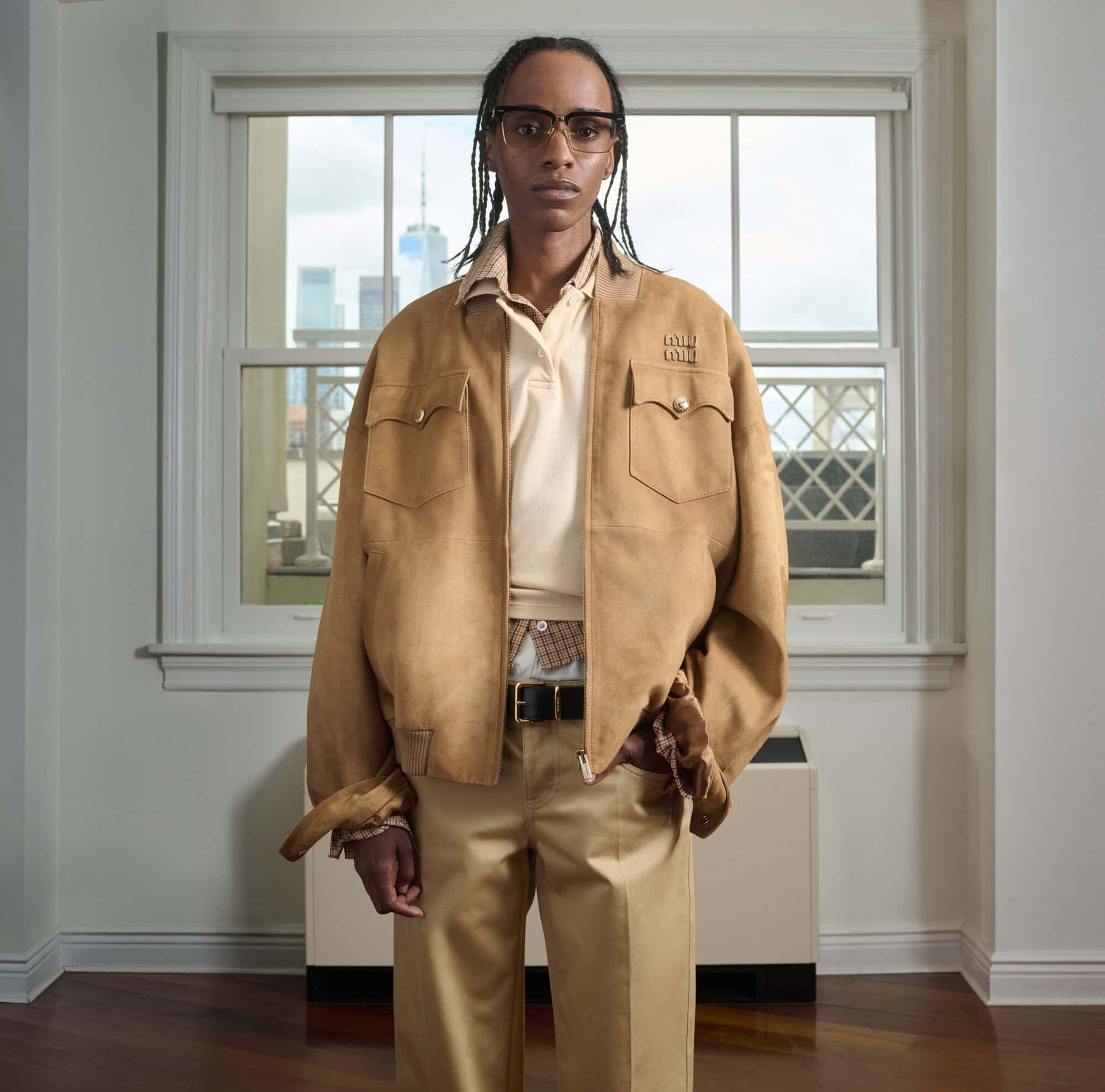 A person with braided hair wearing a tan suede jacket over a beige polo and trousers, standing before a window with a city view.