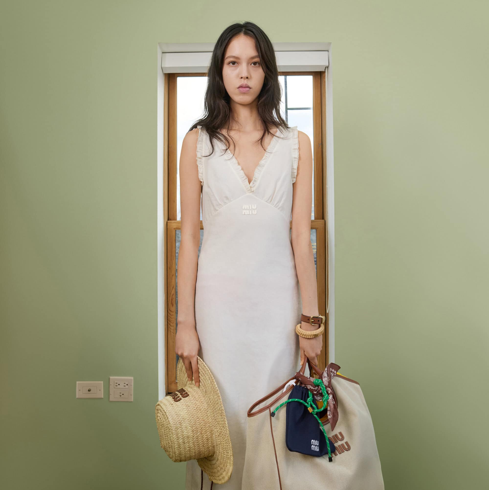 A woman stands in a room with light green walls wearing a white dress, holding a sun hat and a logo branded tote bag.