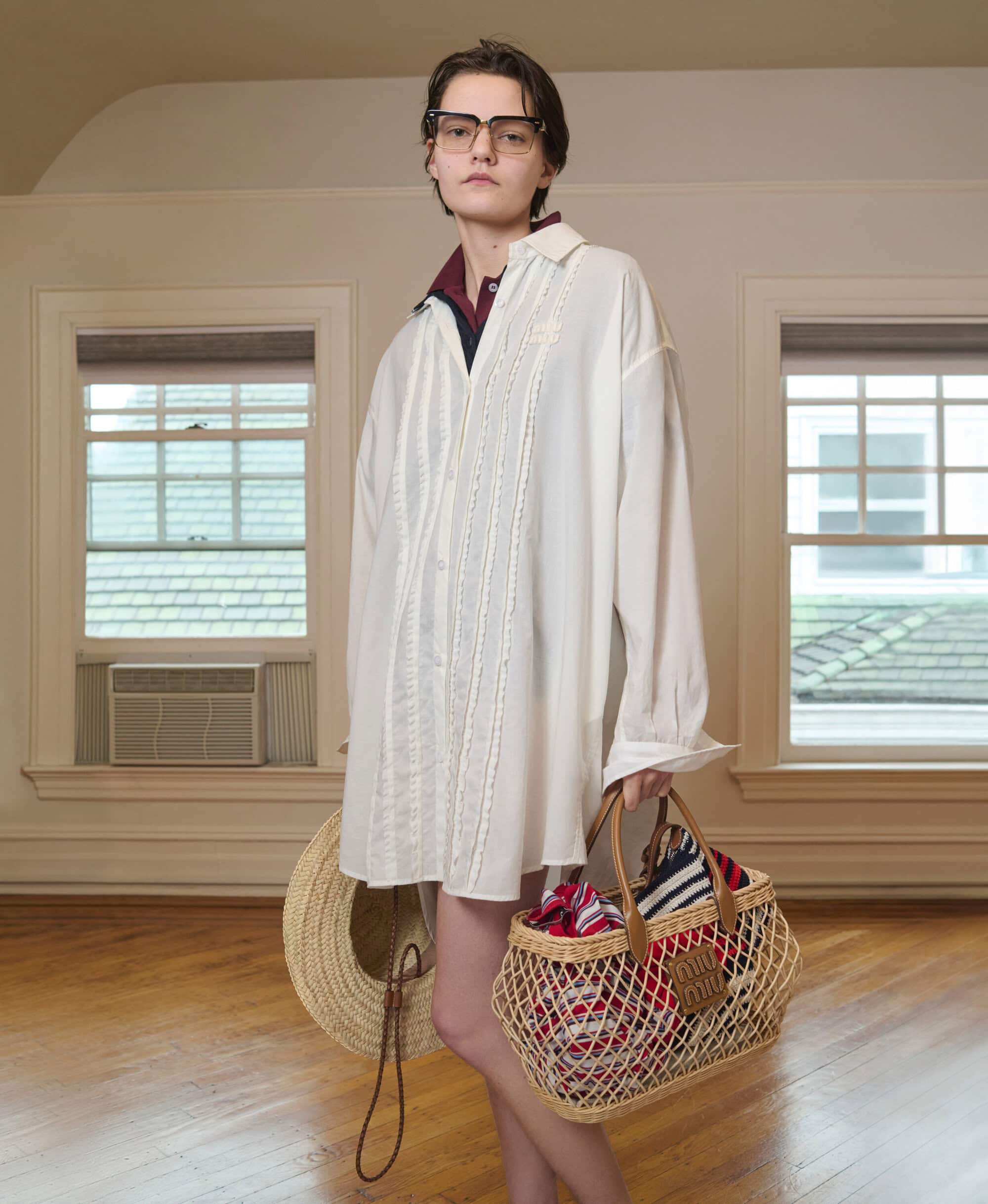 Woman in an oversized white shirt and glasses carries a tote bag and straw hat in a light-filled room with hardwood floors.