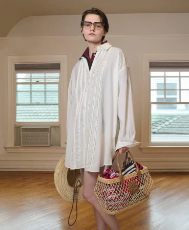 Woman in an oversized white shirt and glasses carries a tote bag and straw hat in a light-filled room with hardwood floors.