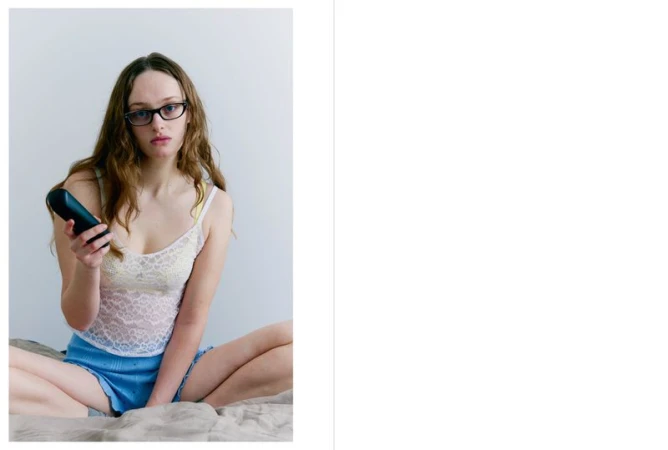Young woman with glasses sits on a bed indoors, holding a remote control, looking toward the camera. She wears a lace top and shorts.