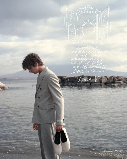 Man in suit stands on beach, holding shoes. Features coastal view, soft light, sketched text overlay, fashion, and seaside mood.