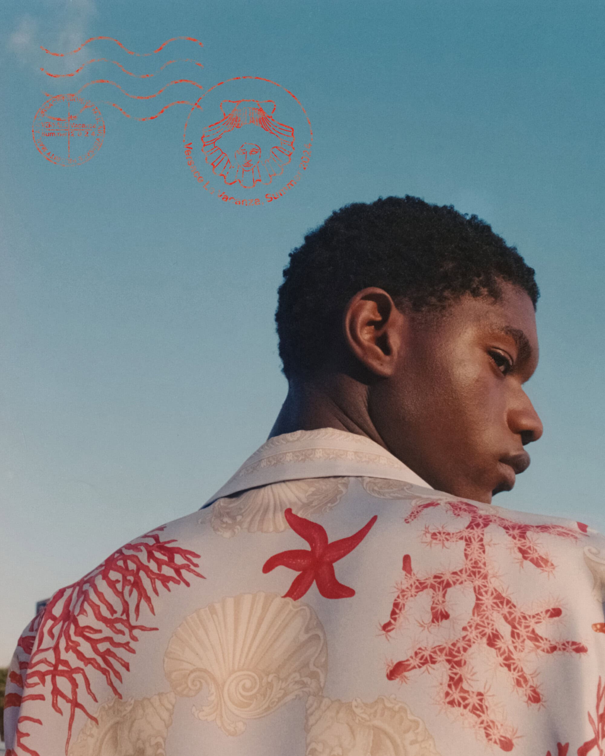Close-up of an adult Black man, seen from the back, wearing a button-down shirt with seashell and coral patterns against a blue sky backdrop.