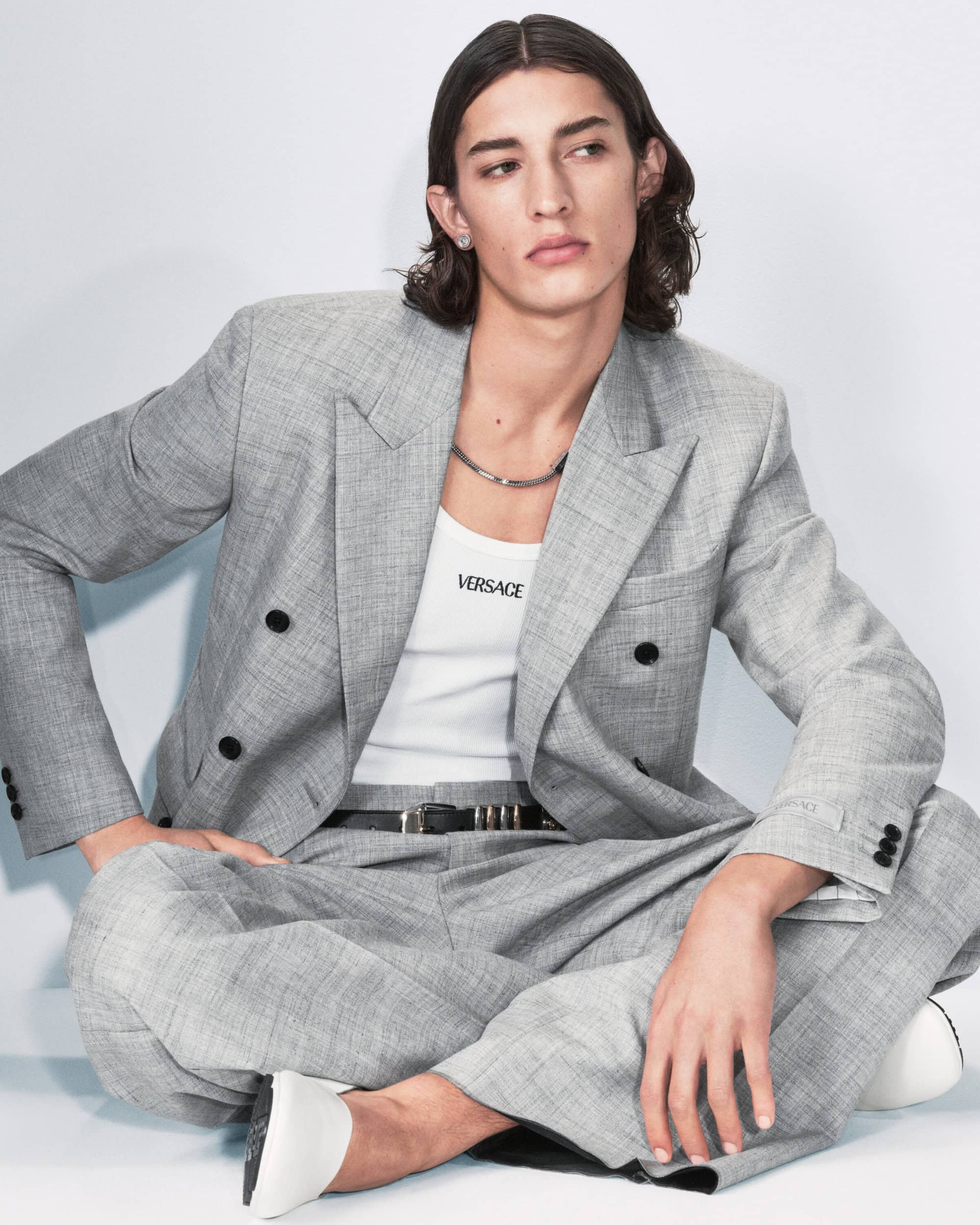 Male model poses in a grey suit and white tank top, seated on the floor in front of a white background, fashion editorial shot.