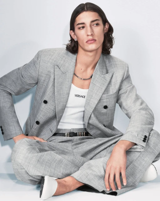 Male model poses in a grey suit and white tank top, seated on the floor in front of a white background, fashion editorial shot.