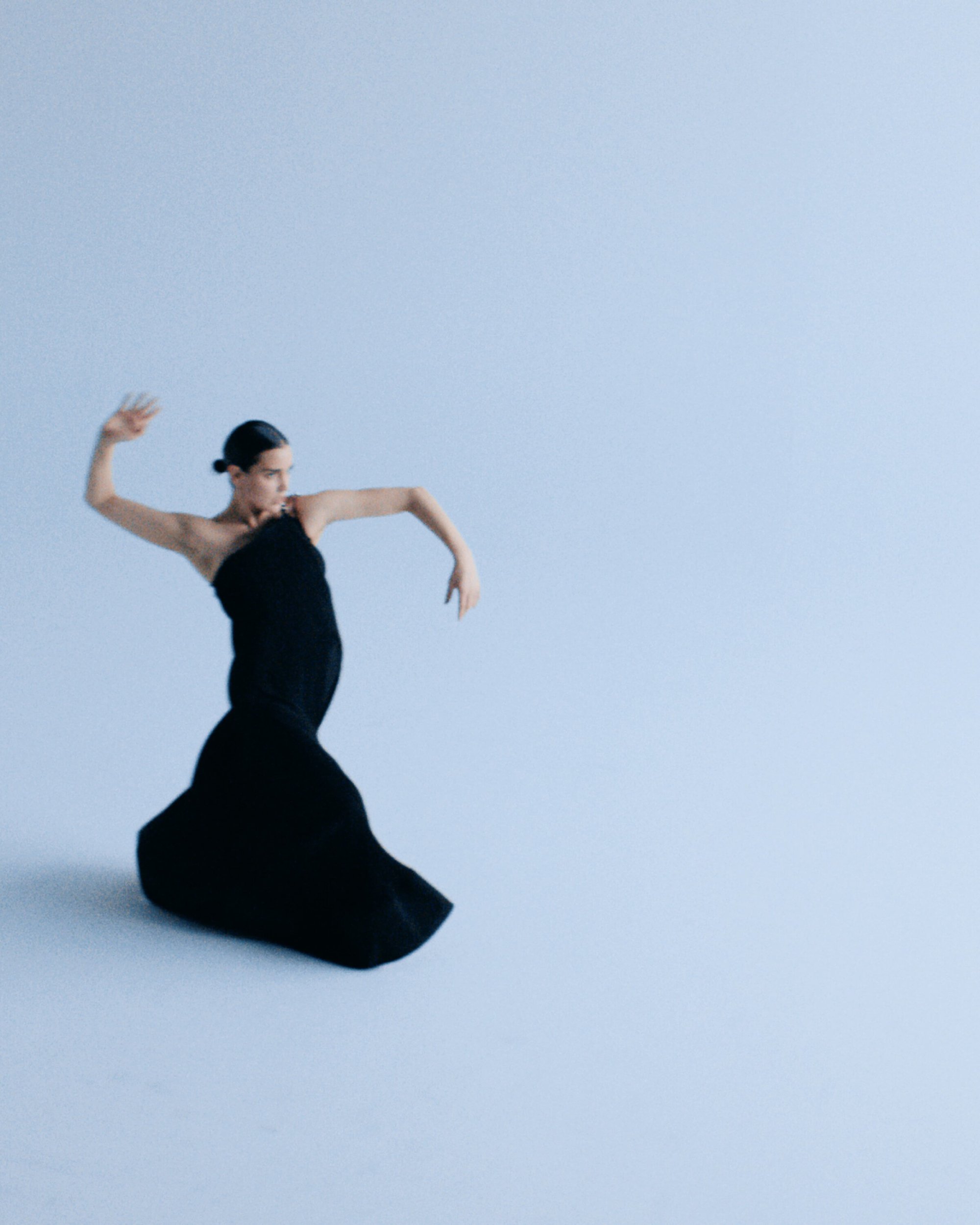 Adult woman in elegant black gown posing gracefully against a light blue backdrop in a studio environment, showcasing fashion and style.