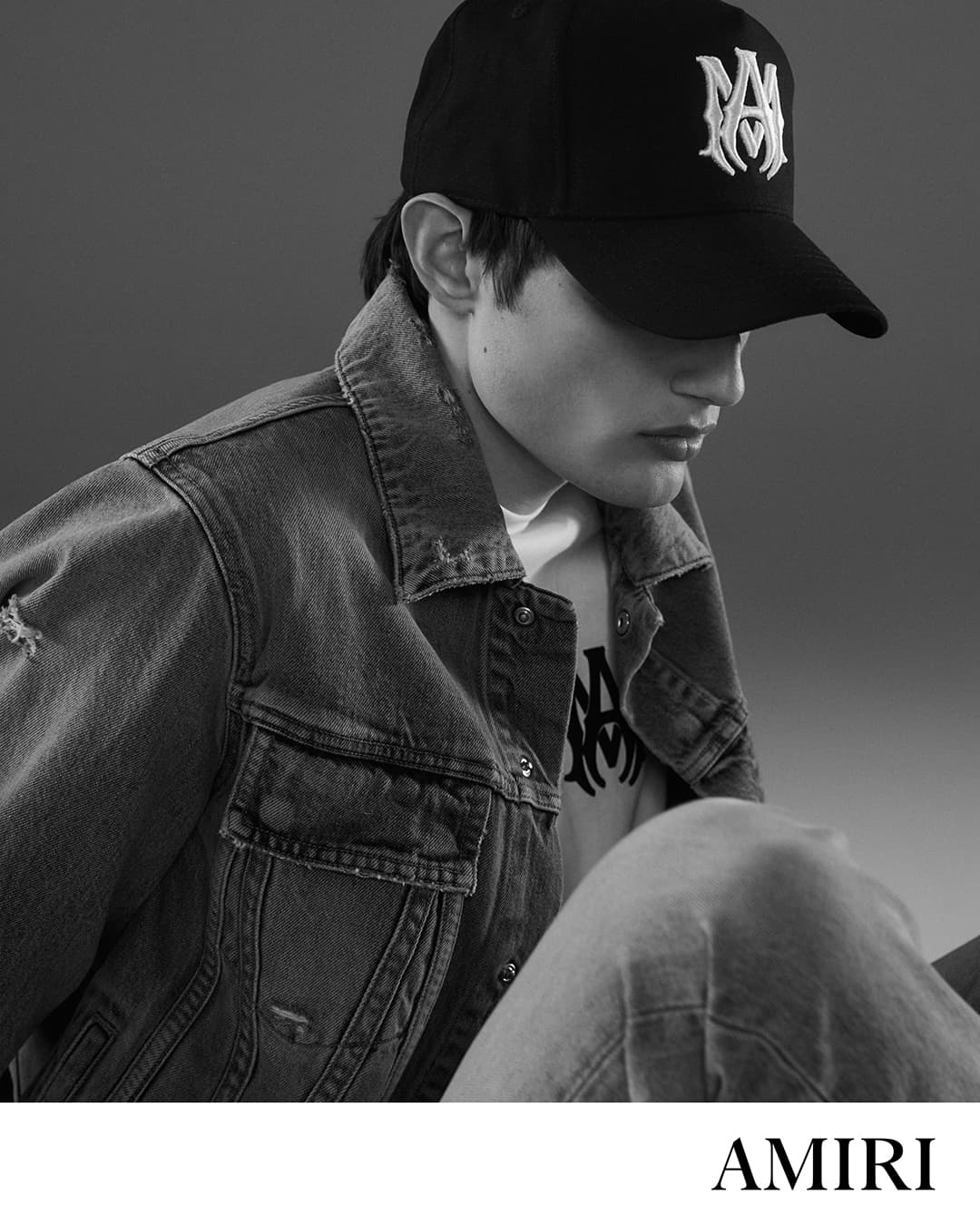 Monochromatic portrait of a male model in a denim jacket and baseball cap against a gray backdrop, fashion photography.