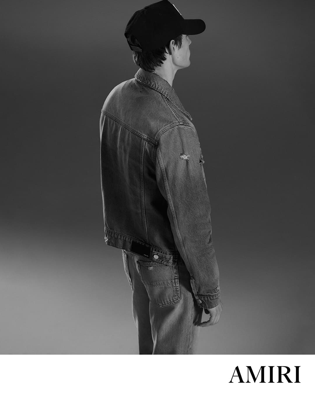Man wearing a denim jacket and baseball cap viewed from behind against a grey backdrop for Amiri Core campaign.