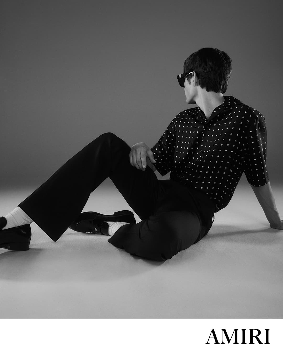 Man seated on a grey seamless floor wearing a polka dot button-down shirt, dark trousers, and sunglasses for Amiri Core campaign