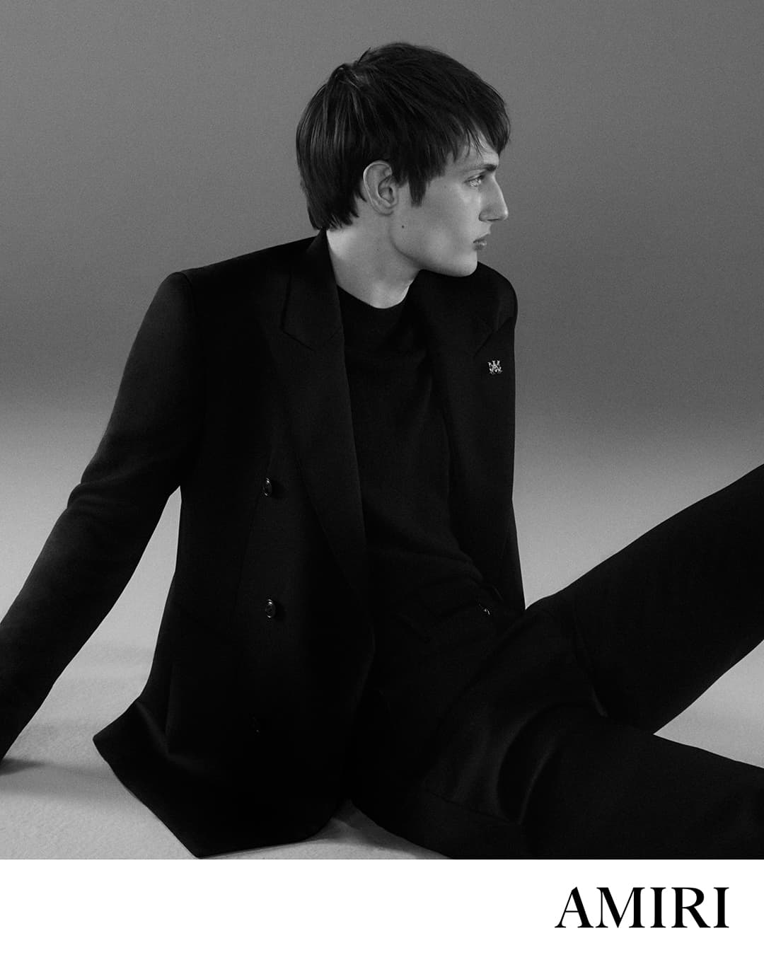 Monochrome fashion shot of a young adult man in a black blazer and pants, seated against a gradient gray background, looking off to the side.