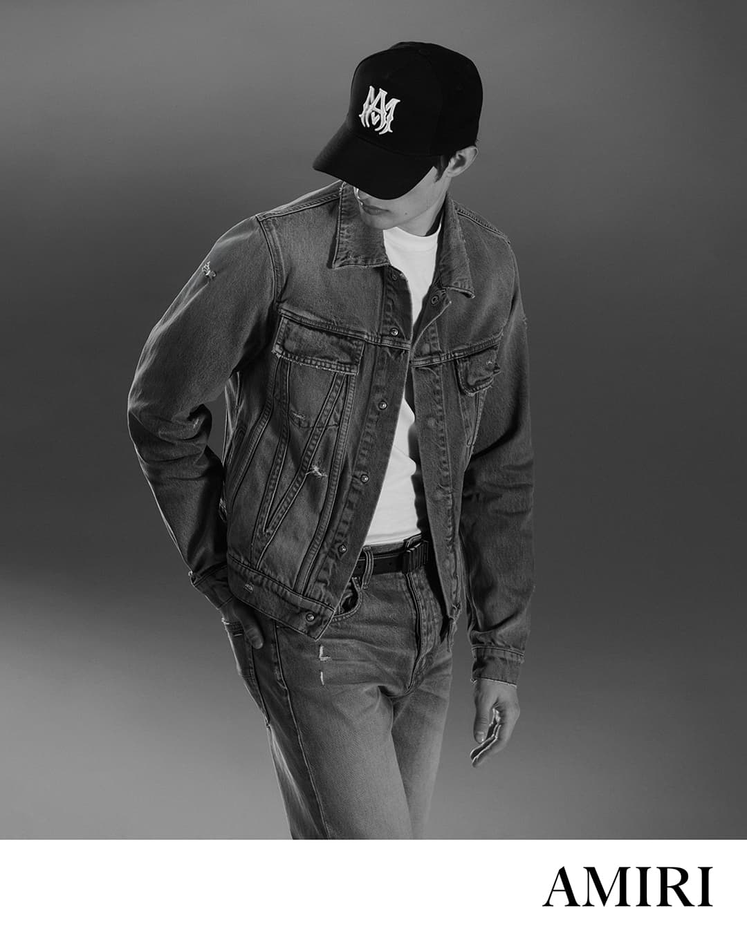 Black and white studio shot of a man wearing a denim jacket, jeans, t-shirt, and baseball cap with a logo, posing in front of a grey backdrop.
