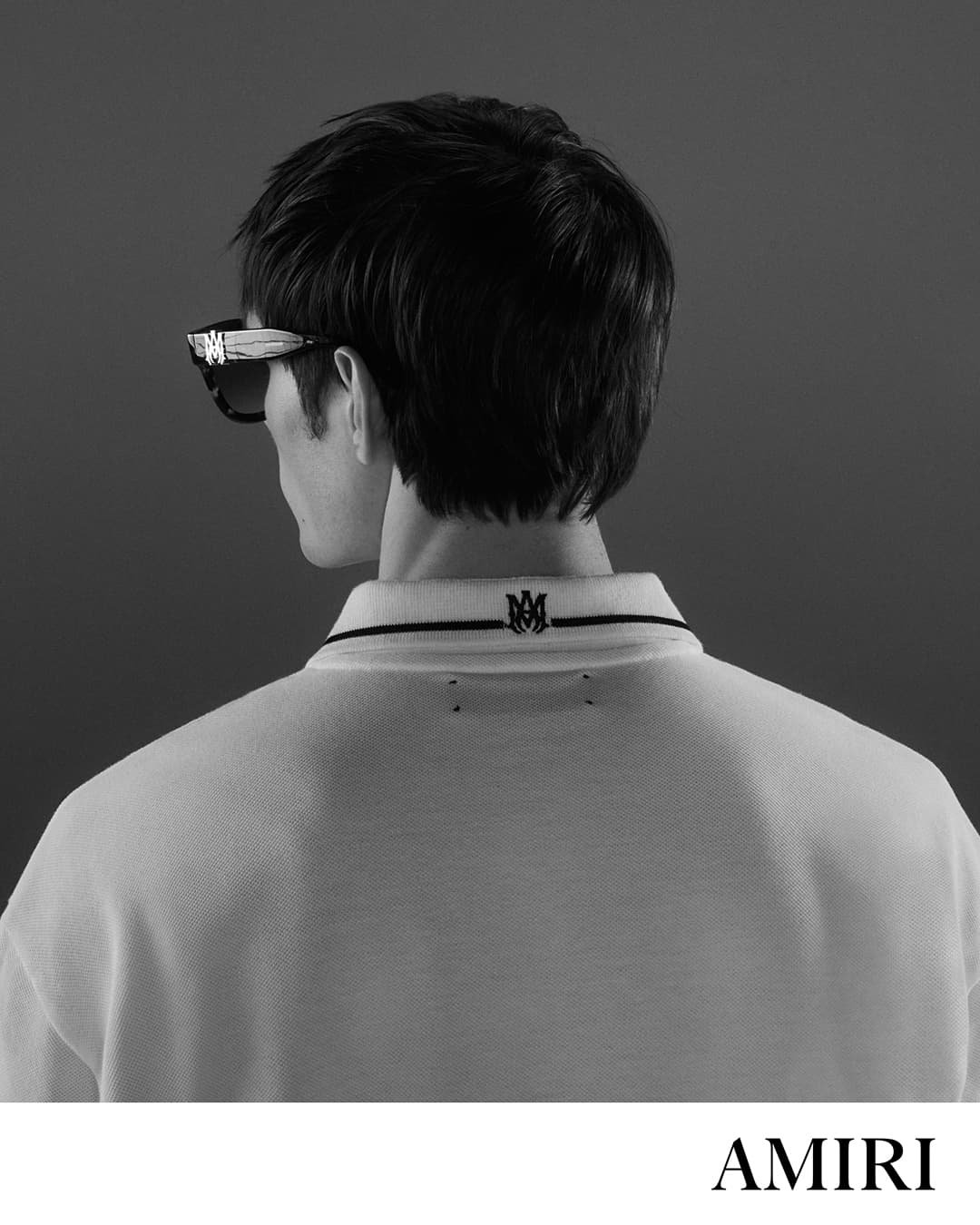 Back of a person's head with black hair wearing sunglasses and a white polo shirt with a black collar, against a grey background.