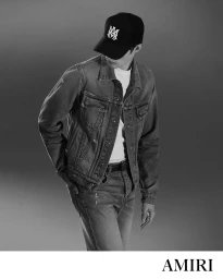 Man wearing a black baseball cap, denim jacket, and jeans against a grey seamless background for Amiri