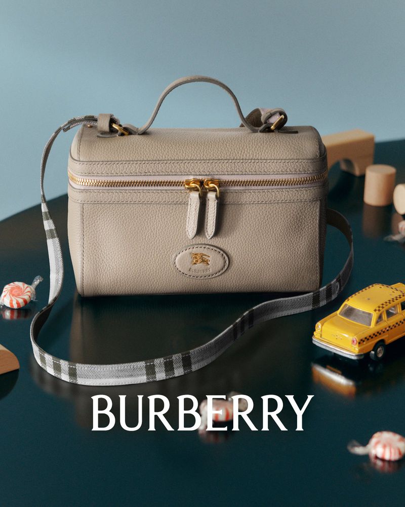 Burberry gray leather vanity case with handle and shoulder strap, set on a dark blue surface with candies and wooden toys.