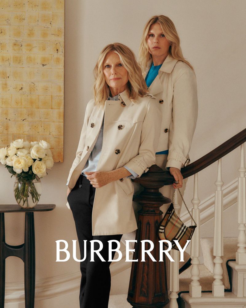 Two women, one middle-aged, one young adult, wearing trench coats and standing on a staircase in a bright indoor setting. Burberry logo.