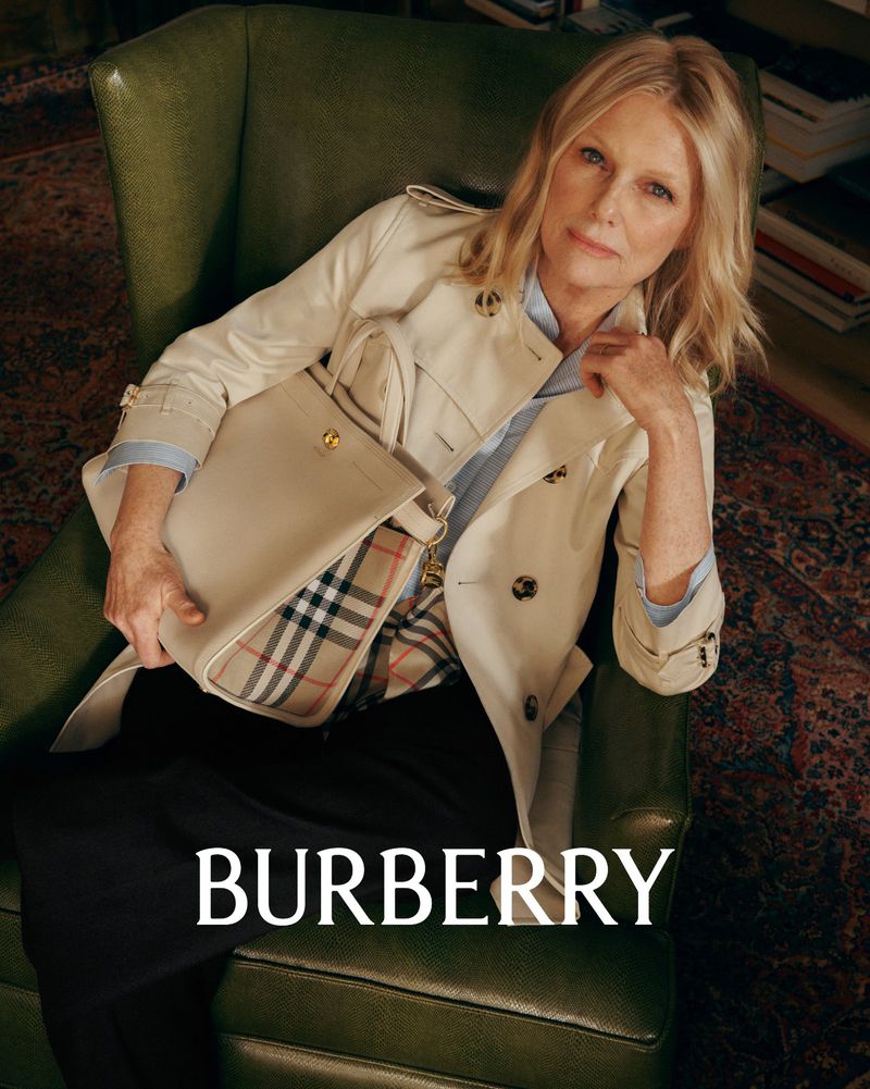 An adult female sits in a green armchair with a Burberry bag, wearing a light-colored trench coat in a bright indoor setting.