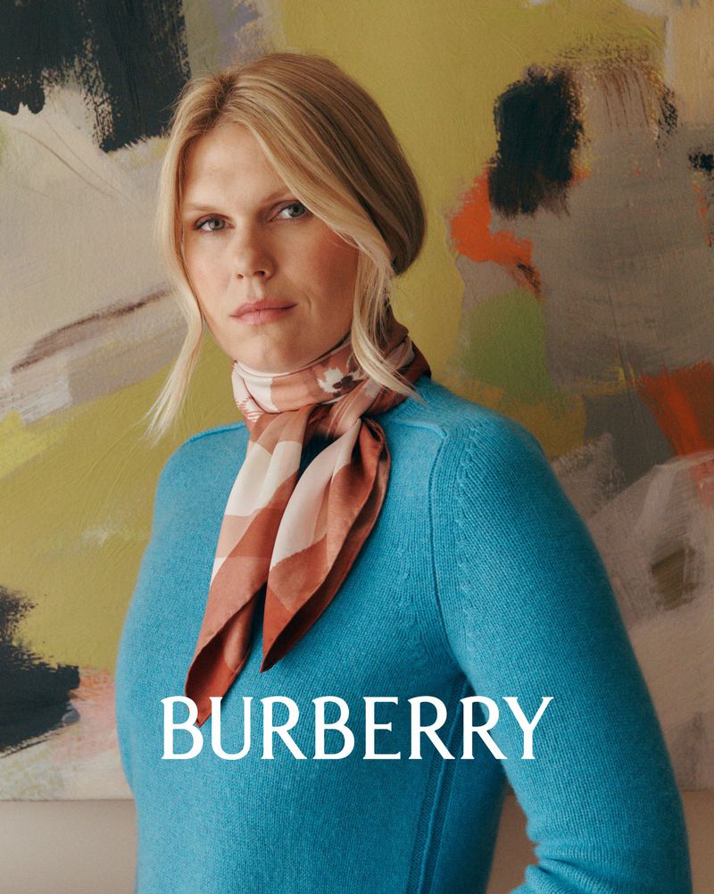 A blonde woman with a tan and brown scarf and turquoise sweater poses in front of abstract painting, promoting Burberry fashion.