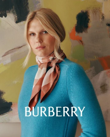 A blonde woman with a tan and brown scarf and turquoise sweater poses in front of abstract painting, promoting Burberry fashion.