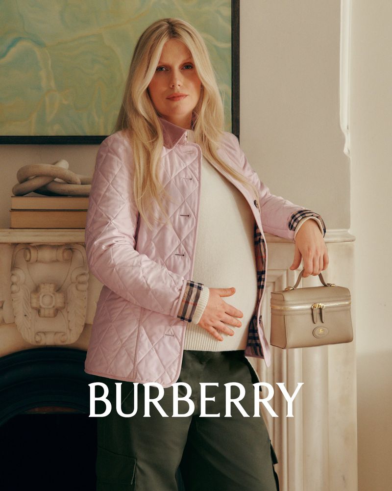 Pregnant woman wearing Burberry jacket and holding a purse poses indoors in front of a light green wall and a fireplace.