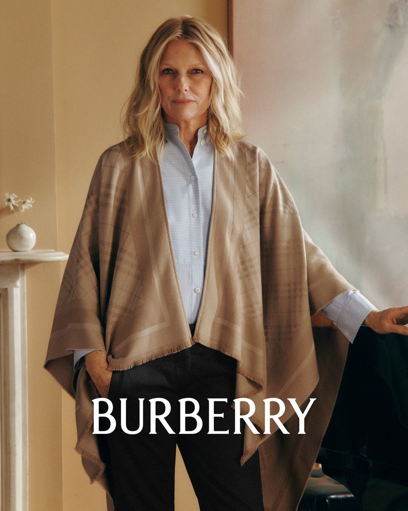 Elegant mature woman with blonde hair wearing a beige Burberry poncho and blue shirt posing indoors in a fashion portrait with neutral background.