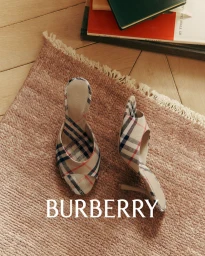 Pair of Burberry plaid check high heel mules resting on a textured pink rug next to books on a wooden floor