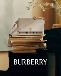 A white Burberry handbag with plaid trim rests on a tall stack of vintage books atop a black piano indoors.