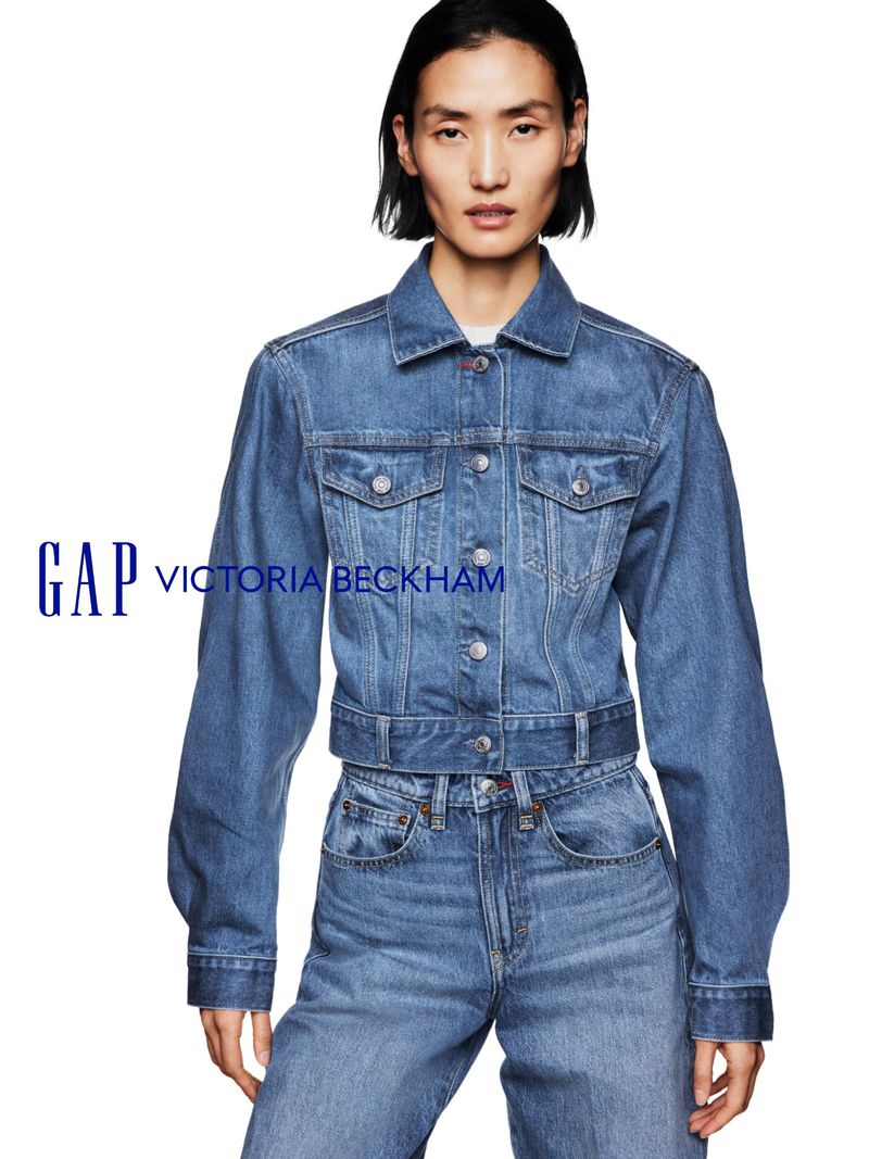 A woman in a denim jacket and jeans poses against a white backdrop, showcasing a GAP x Victoria Beckham collaboration with the logo.