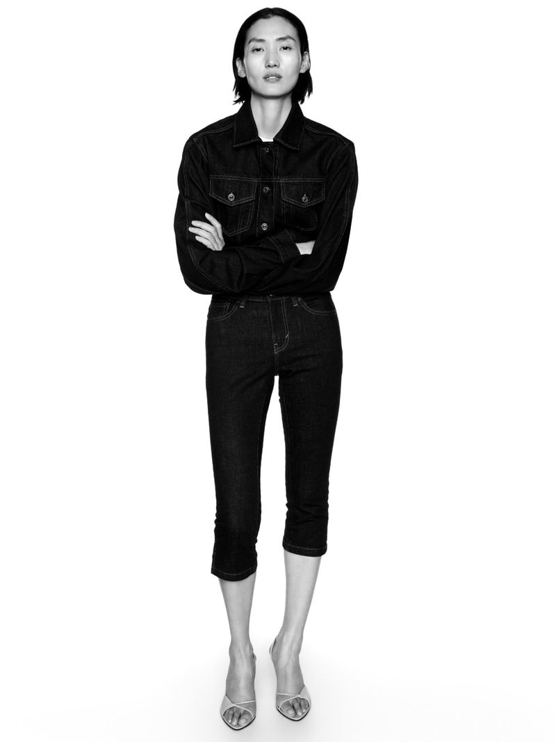 Full-length black and white studio portrait of an Asian woman in dark denim jacket and cropped pants, arms crossed, on white background.